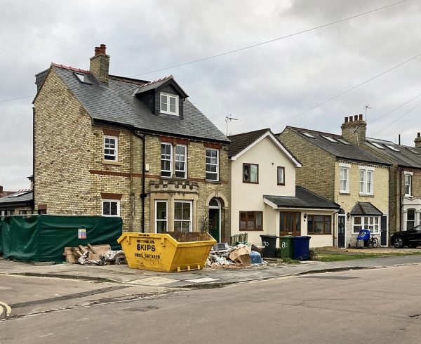 A brick house with a new loft dormer and rendered side extension under construction, with a skip and hoarding outside